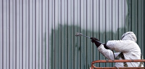 A commercial painter on an industrial lift spray painting a steel exterior wall or duct.
