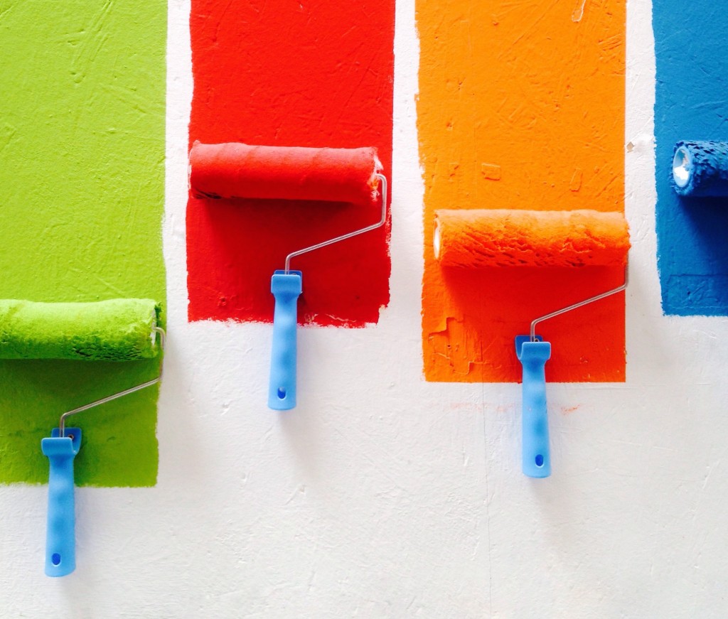four paint rollers coating a while wall with different primary colours