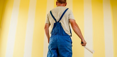 A professional painter stands in front of a yellow walll, painting the wall white in stripes, he is holding a paint roller